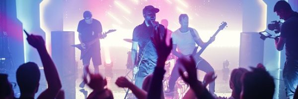 Rock Band with Guitarists and Drummer Performing at a Concert in a Night Club. Front Row Crowd is Partying. Silhouettes of Fans Raise Hands in Front of Bright Colorful Strobing Lights on Stage.